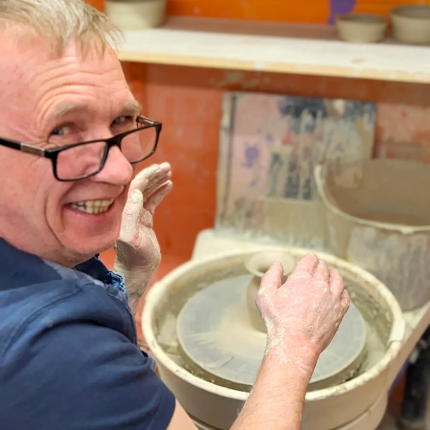 Student grinning with clay-covered hands at a pottery wheel